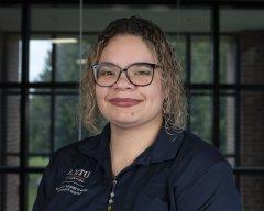 Mercedes Steele, in glasses and a blue Utica University polo shirt, stands in front of a window and smiles.